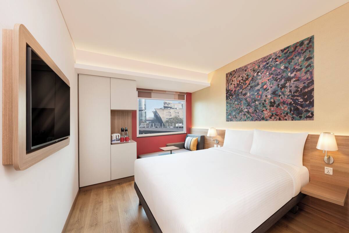 A modern hotel room with a large bed, a flat-screen TV mounted on the wall, a window with a city view, and a colorful abstract painting above the bed.