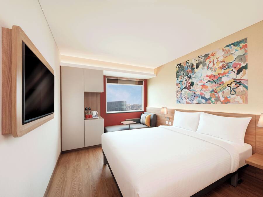 A hotel room with a bed, a TV mounted on the wall, a window, a painting, and a desk with a chair.