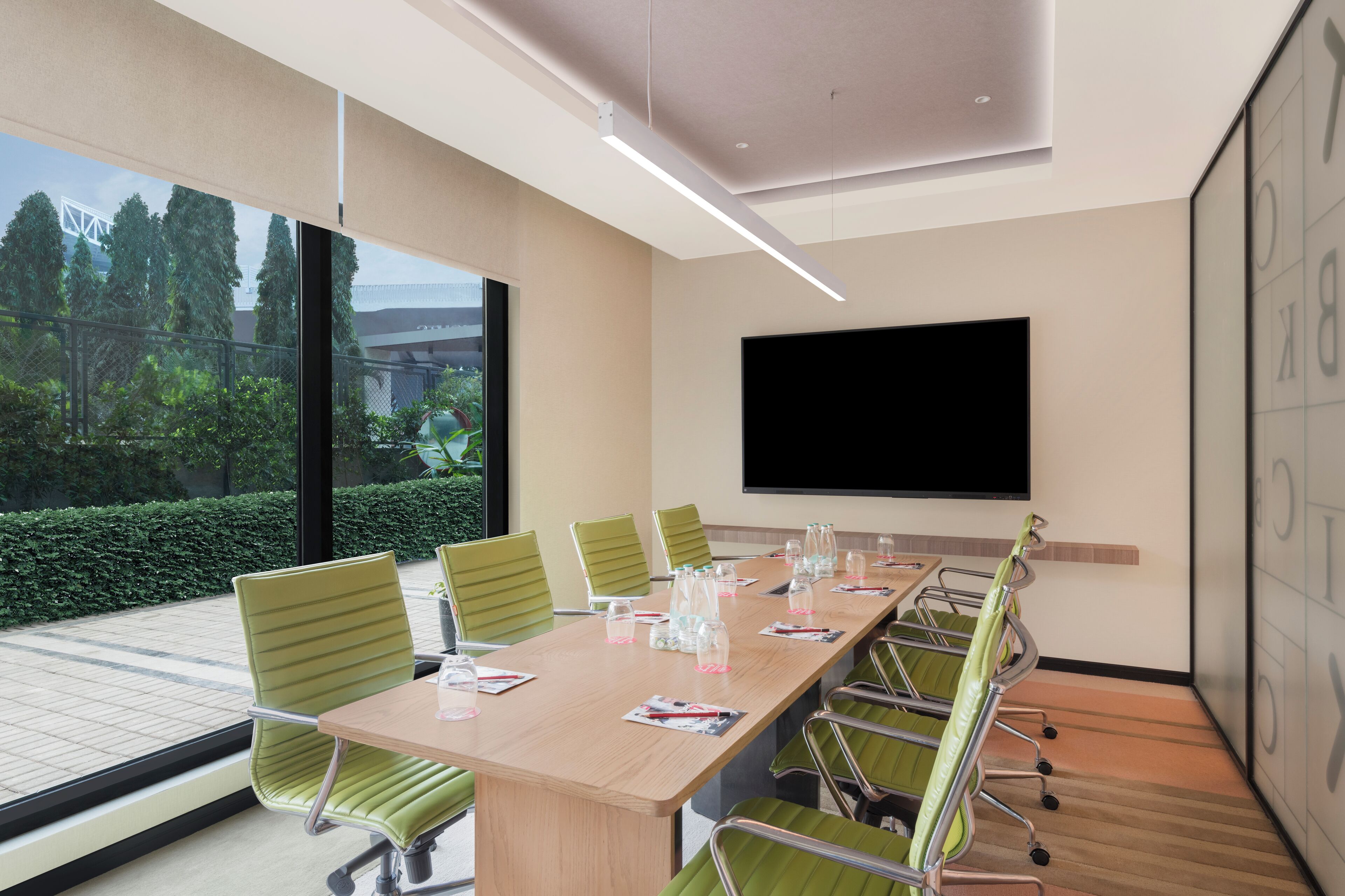 Un salón de conferencias con una larga mesa, sillas verdes, una pantalla grande y vistas al exterior con follaje.