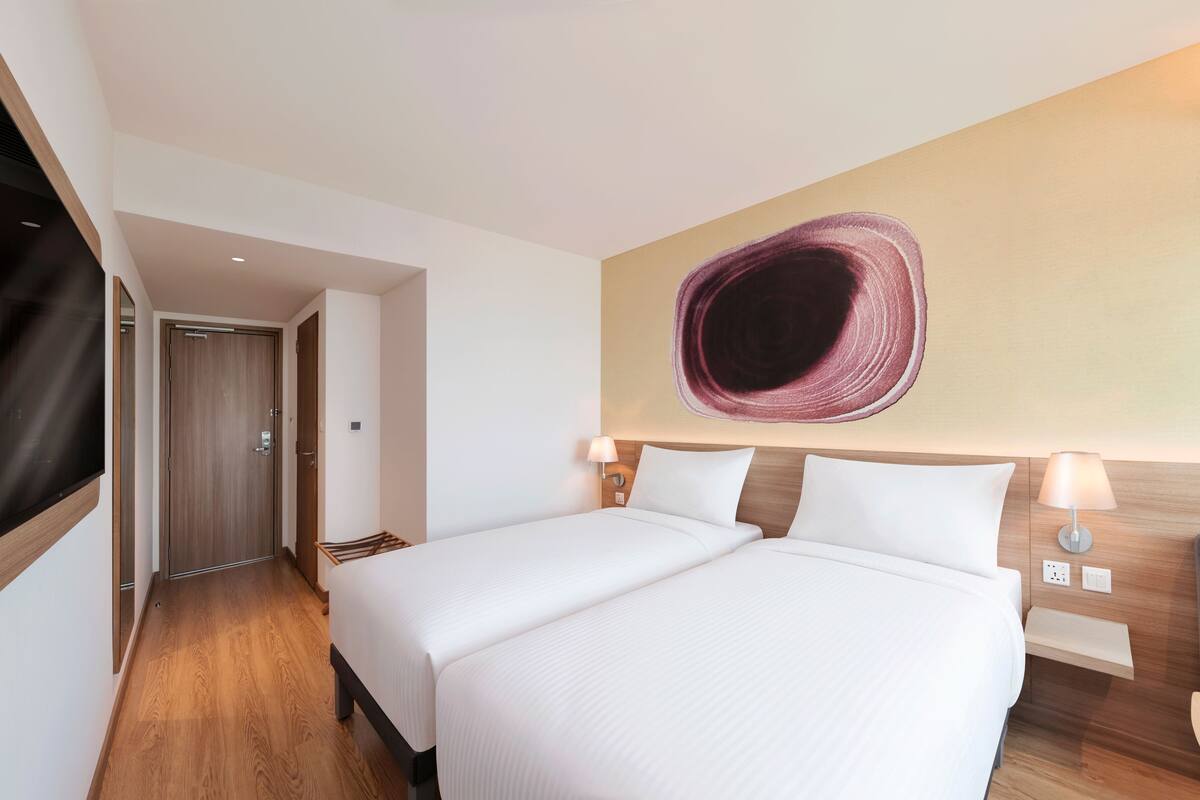 A hotel room with two beds, a wooden floor, a wall-mounted TV, and a modern abstract painting.