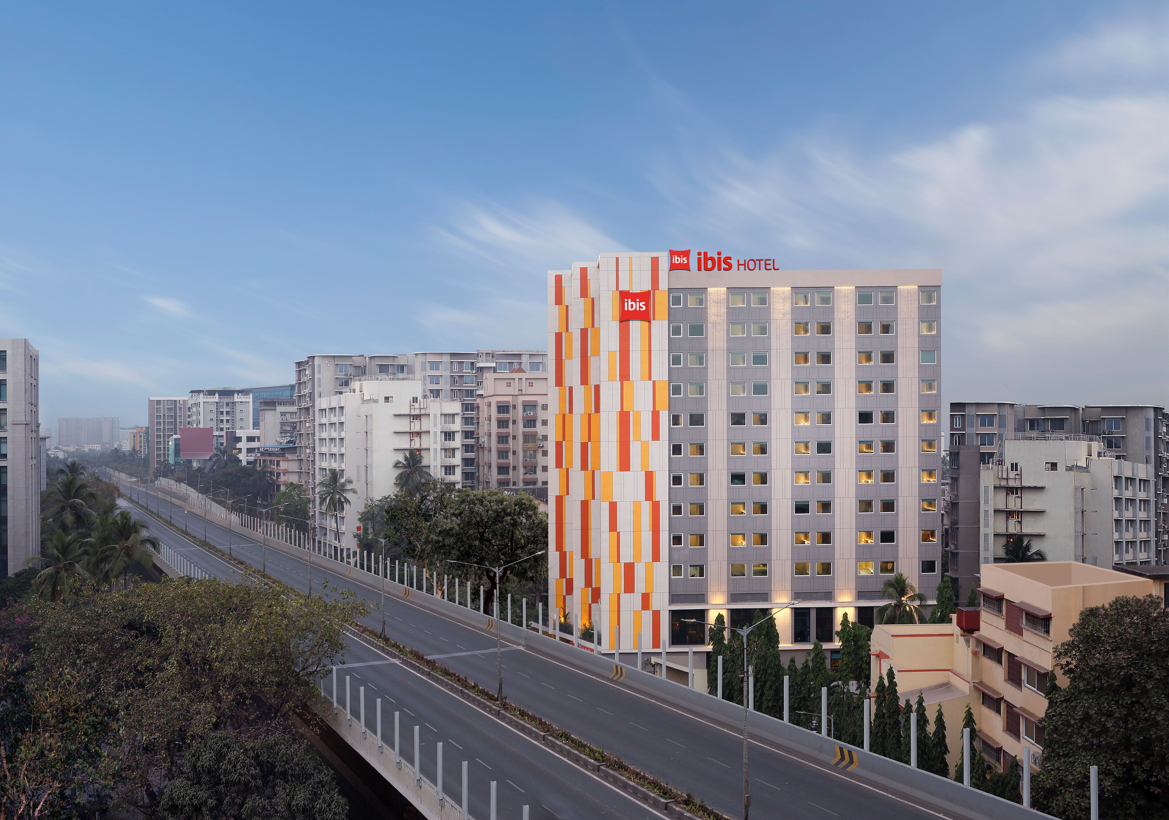 A modern hotel with a distinctive orange and white facade, multiple floors, and a well-maintained surrounding area with trees and a clear sky.