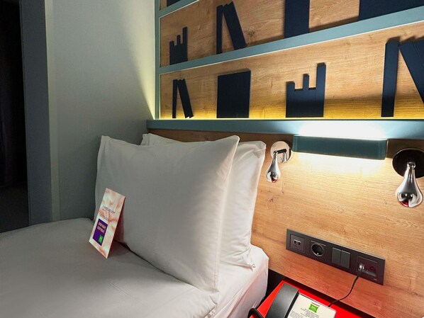 Habitación de hotel con cama, cabecera de madera con letras recortadas, mesita de noche con lámpara y enchufe.