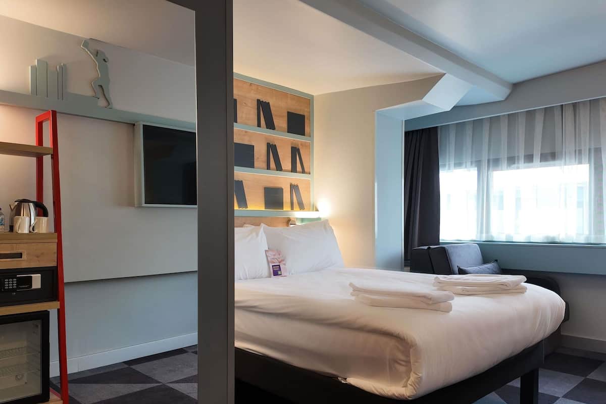 A hotel room with a bed, a desk with a kettle, a shelf with books, and a window with curtains.