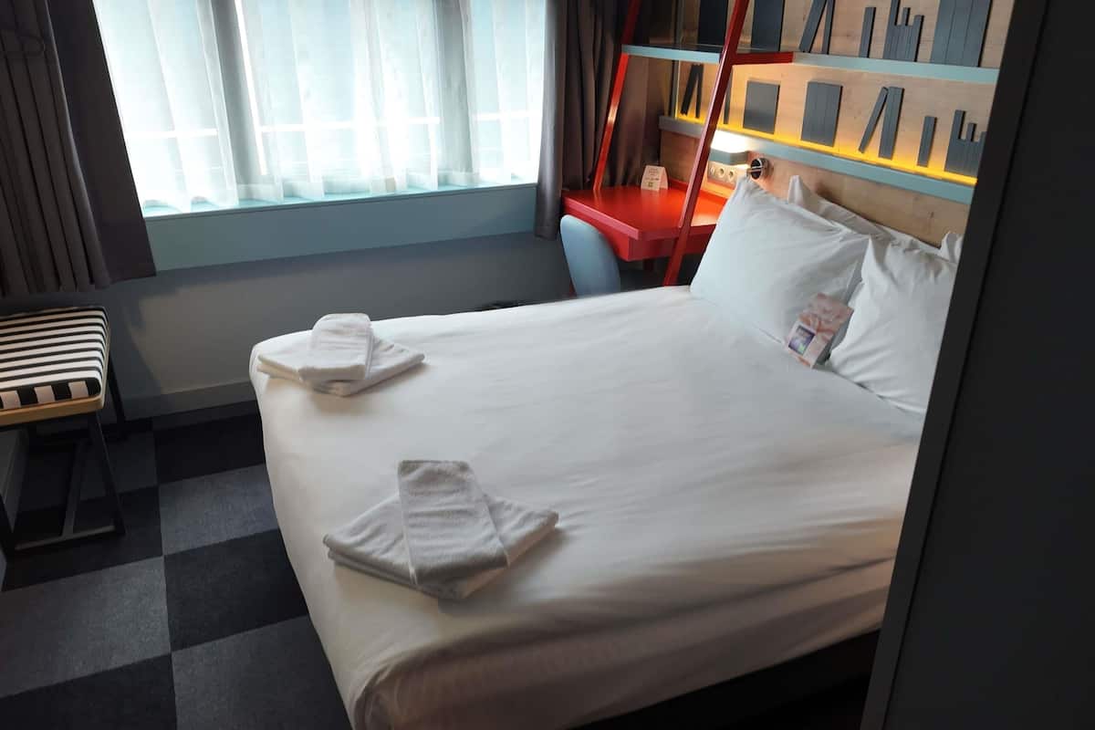 A hotel room with a neatly made bed, a desk, and a window with curtains.