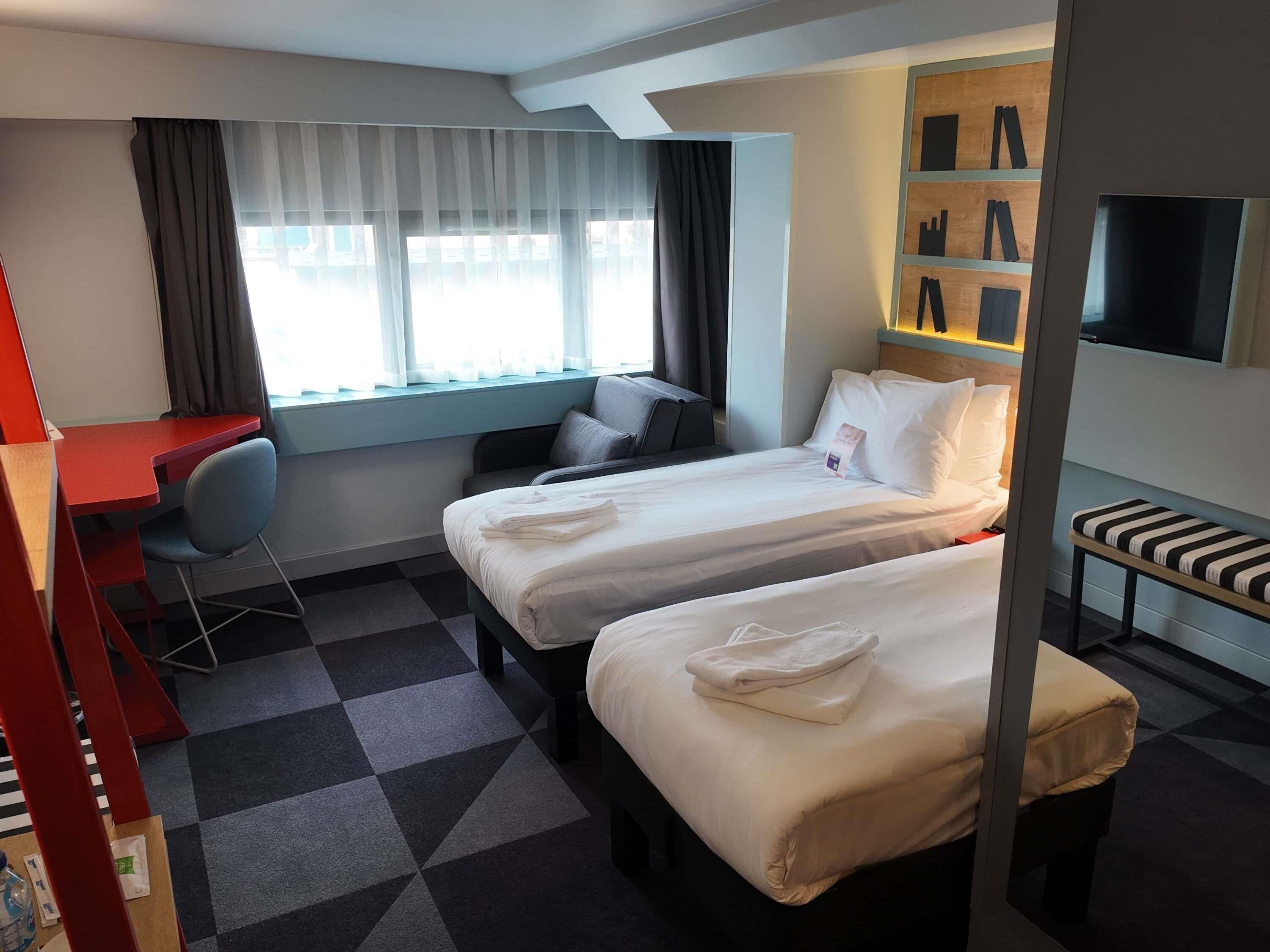 A hotel room with two beds, a desk, a chair, a TV, and a window with curtains.
