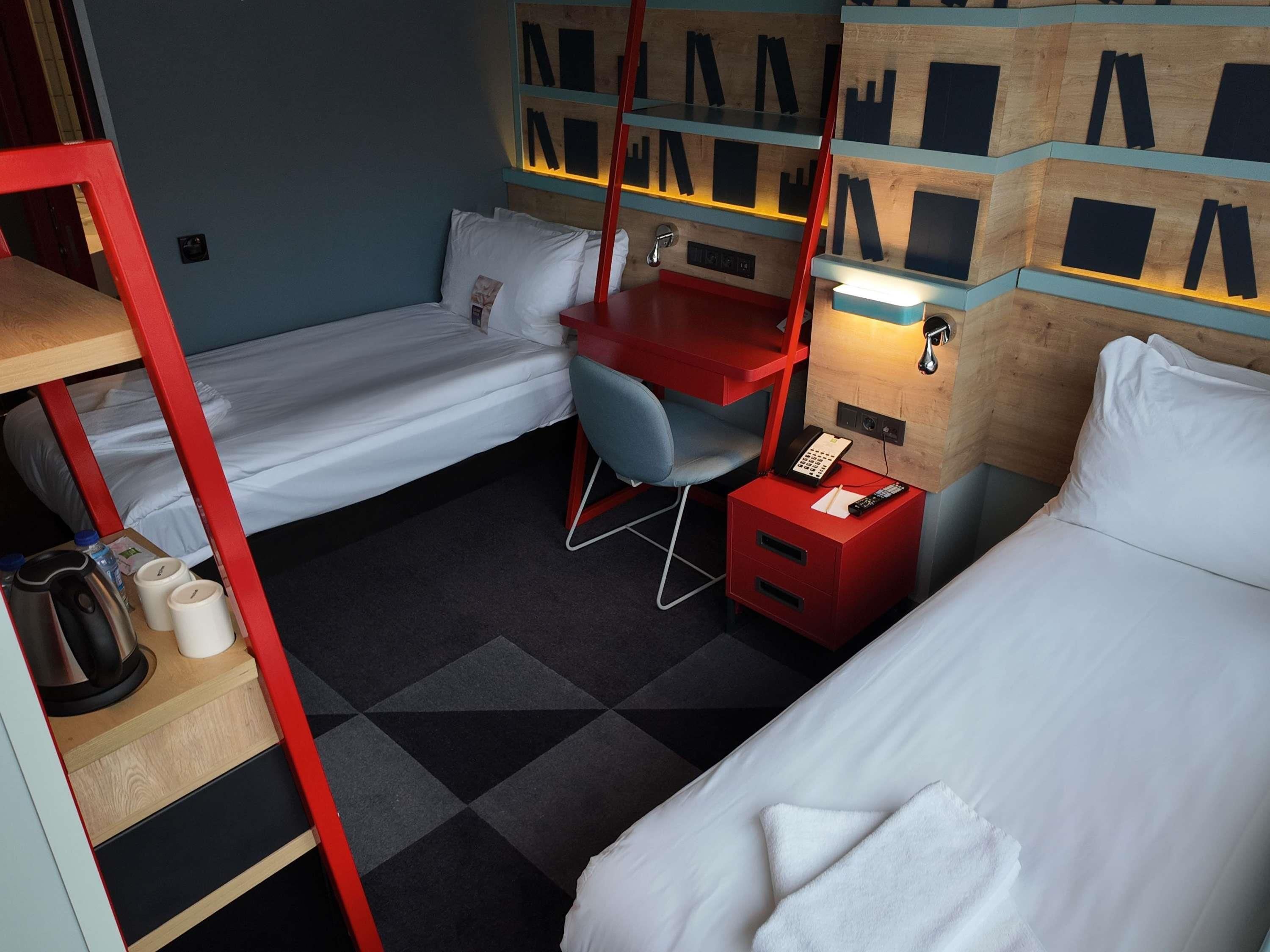 Hotellihuone, jossa on kaksi erillistä sänkyä, työpöytä tuolilla, punainen yöpöytä, jossa on puhelin, ja hylly, jossa on kehystettyjä valokuvia.
