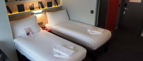 A hotel room with two single beds, a nightstand with a lamp, a wall-mounted shelf with black objects, a red door, and a black and grey patterned carpet.