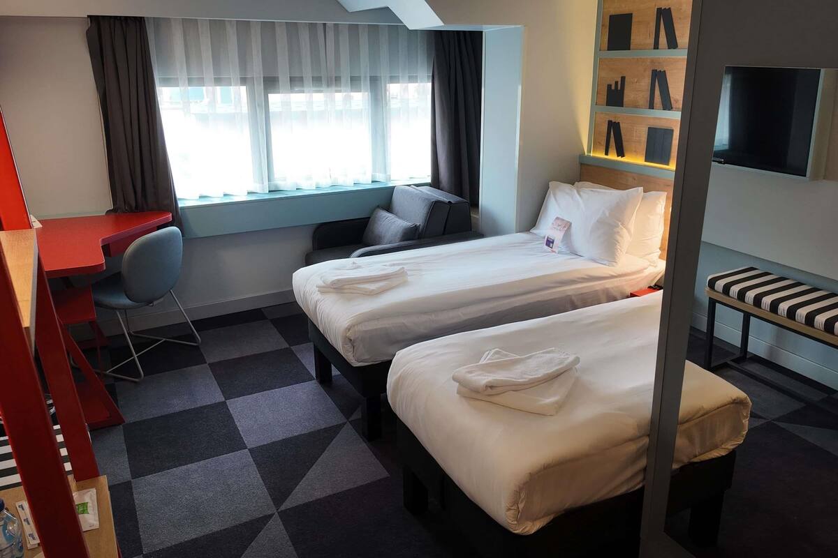 A hotel room with two beds, a desk, a chair, a TV, and a window with curtains.