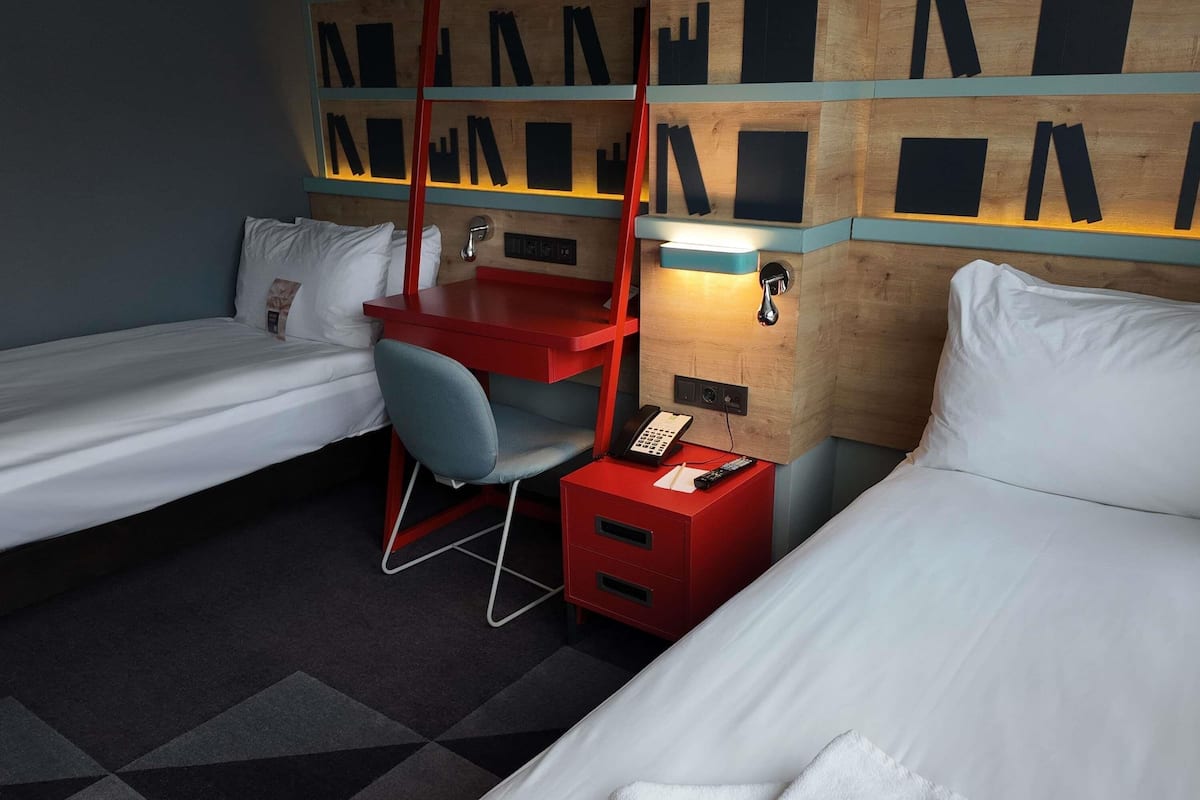 A hotel room with two single beds, a desk with a chair, a red nightstand with a phone and remote, and a wall with wooden panels and black shapes.