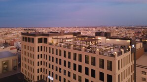 Un hotel de varios pisos, una terraza en la azotea con asientos al aire libre y vistas a la ciudad al fondo.