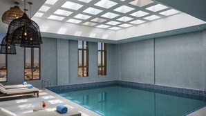 Una piscina interior moderna con agua cristalina azul, grandes ventanales y zonas de descanso con tumbonas.