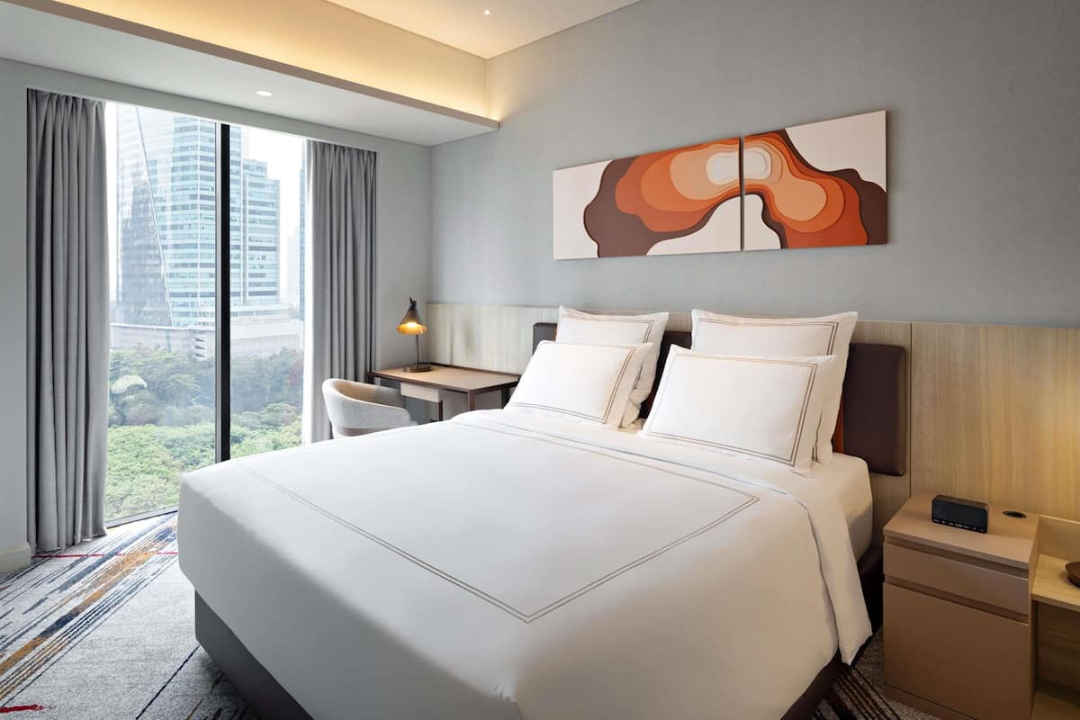 A hotel room with a large bed, a desk, a chair, a nightstand, and a view of the cityscape.