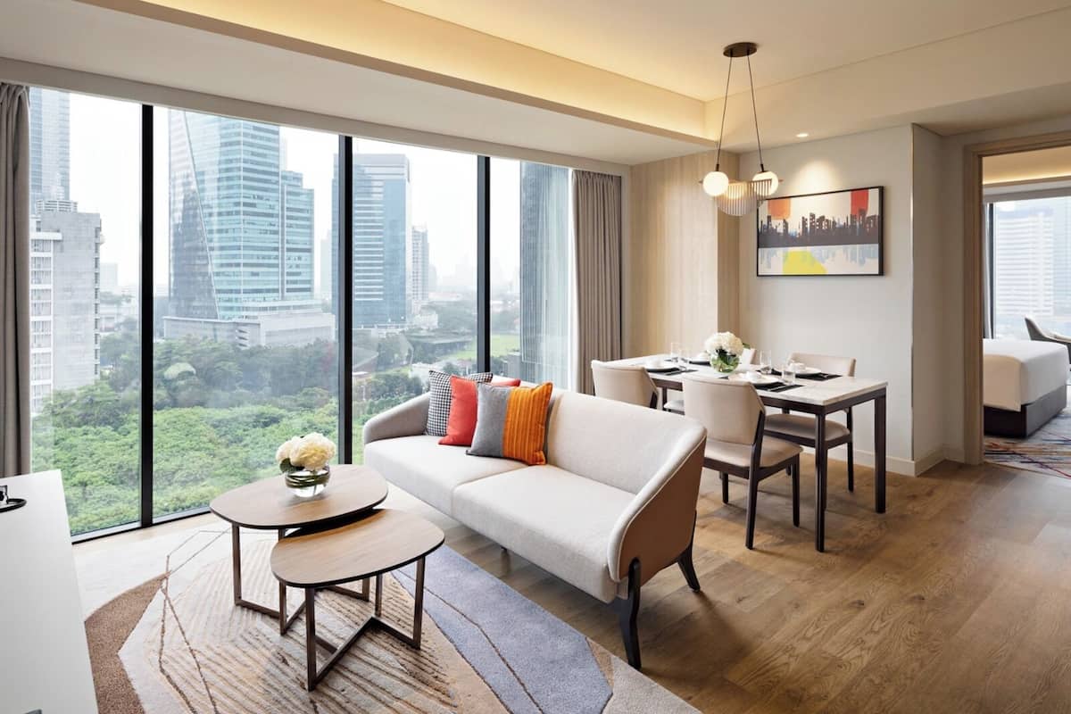 A modern hotel room with a sofa, dining table, and chairs, large windows with city views, and a cityscape painting on the wall.