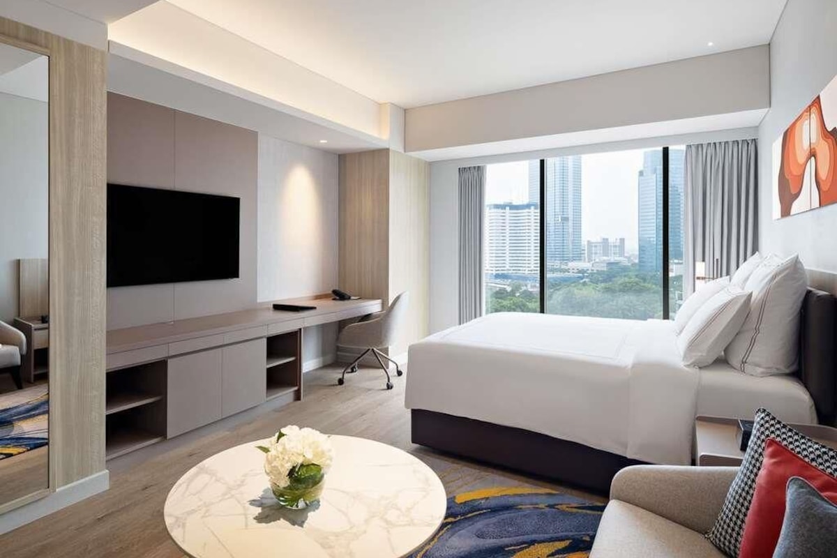 Une chambre d’hôtel moderne dotée d’un grand lit, d’une télévision à écran plat, d’un bureau et offrant une vue sur le paysage urbain.