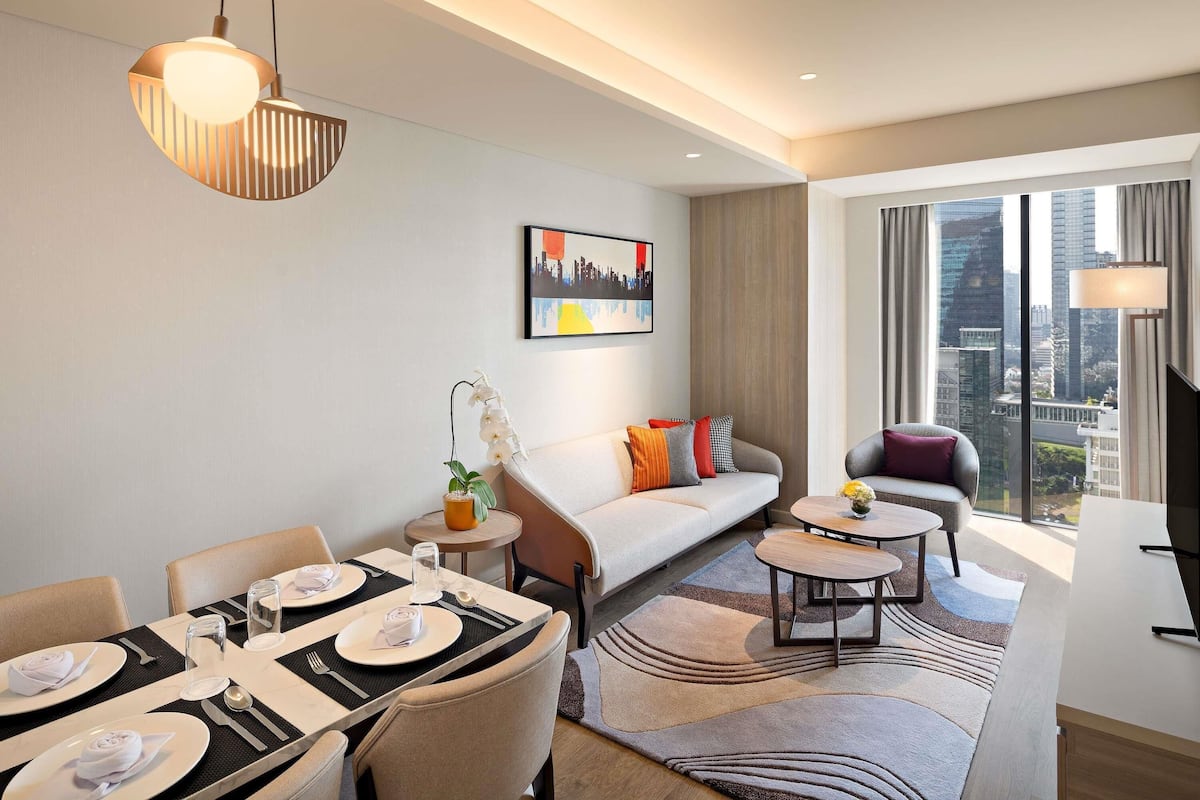 A modern hotel room with a dining area, a sofa, a TV, and a city view.