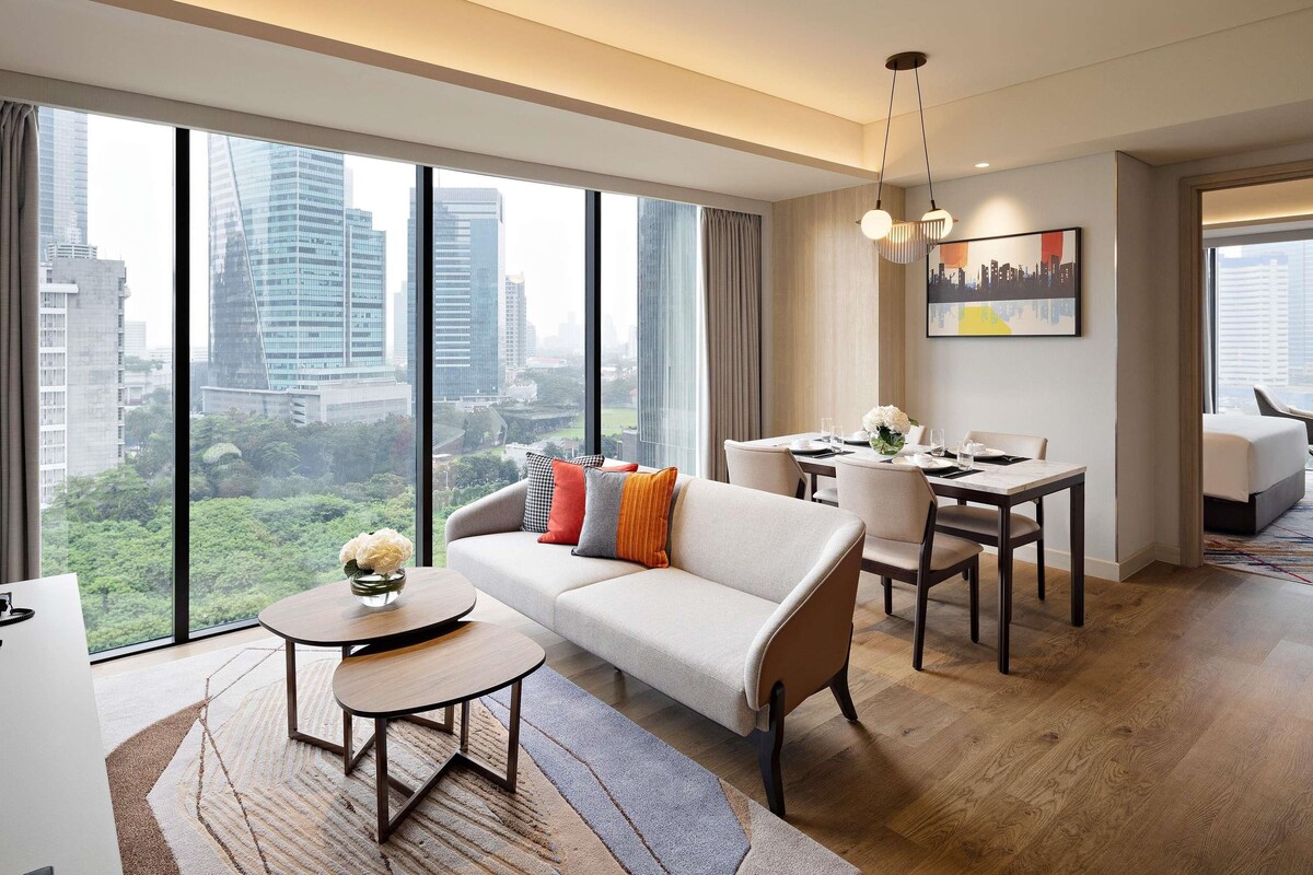A modern hotel room with a sofa, dining table, and chairs, large windows with a city view, and a painting on the wall.