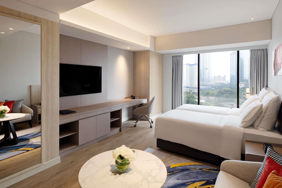 A modern hotel room with a large bed, a desk with a flat-screen TV, a seating area with a sofa and armchairs, and a city view through large windows.