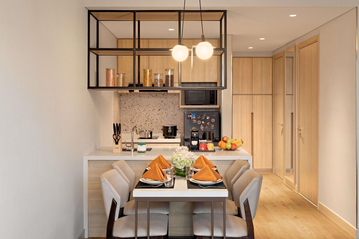 A modern kitchen with a dining area, featuring a central island, overhead lighting, and wooden cabinetry.