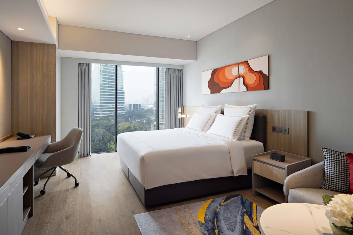A modern hotel room with a large bed, a desk, and a chair. There is a view of the city through large windows.