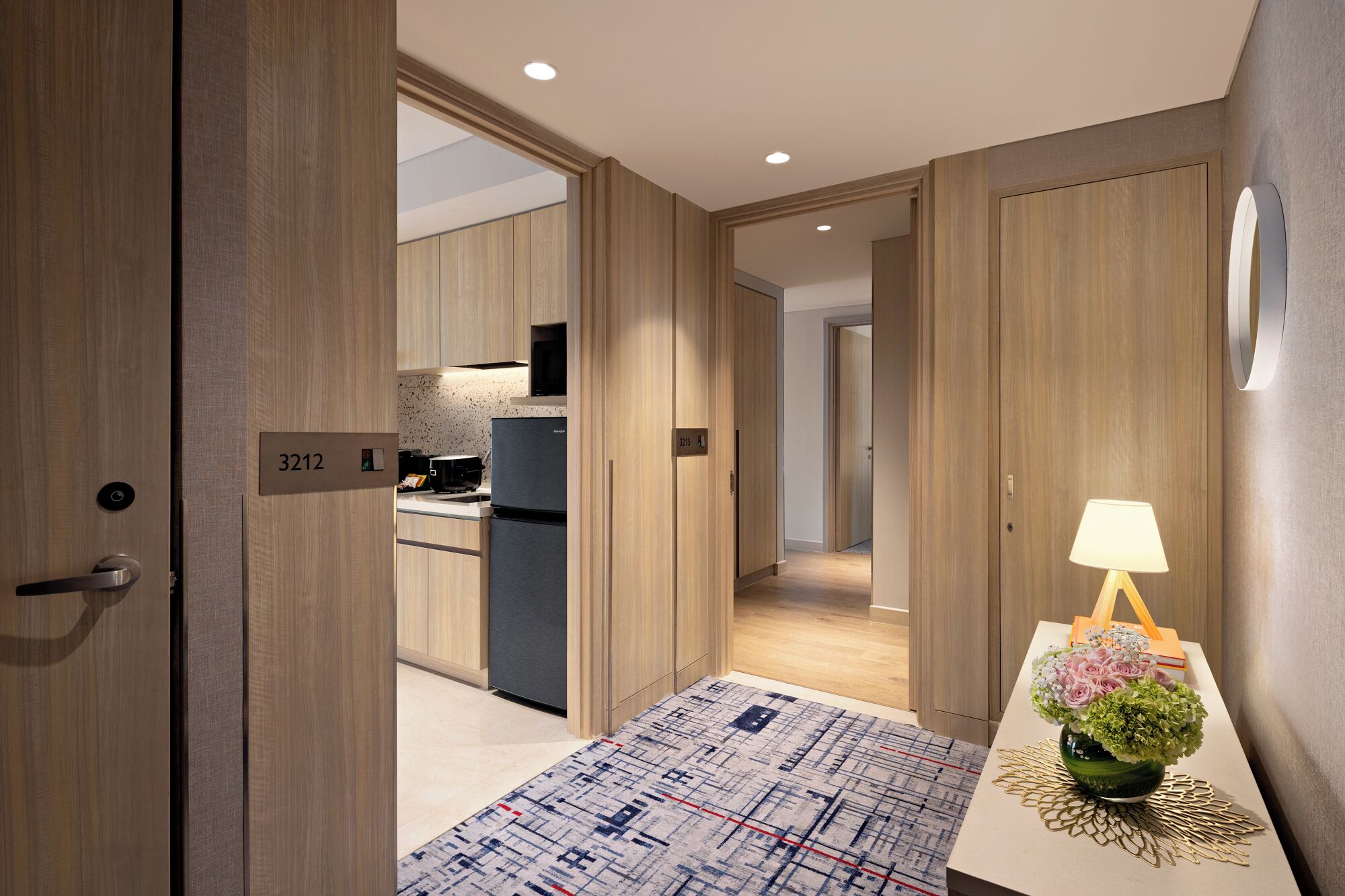 Een moderne hotelkamer met houten vloeren, een kitchenette en een nachtkastje met lamp en bloemen.