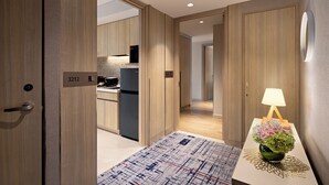 Una habitación de hotel moderna con suelo de madera, zona de cocina y mesita de noche con lámpara y flores.
