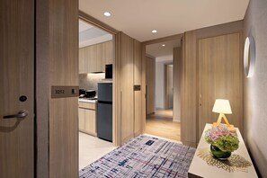Una habitación de hotel moderna con suelo de madera, zona de cocina y mesita de noche con lámpara y flores.