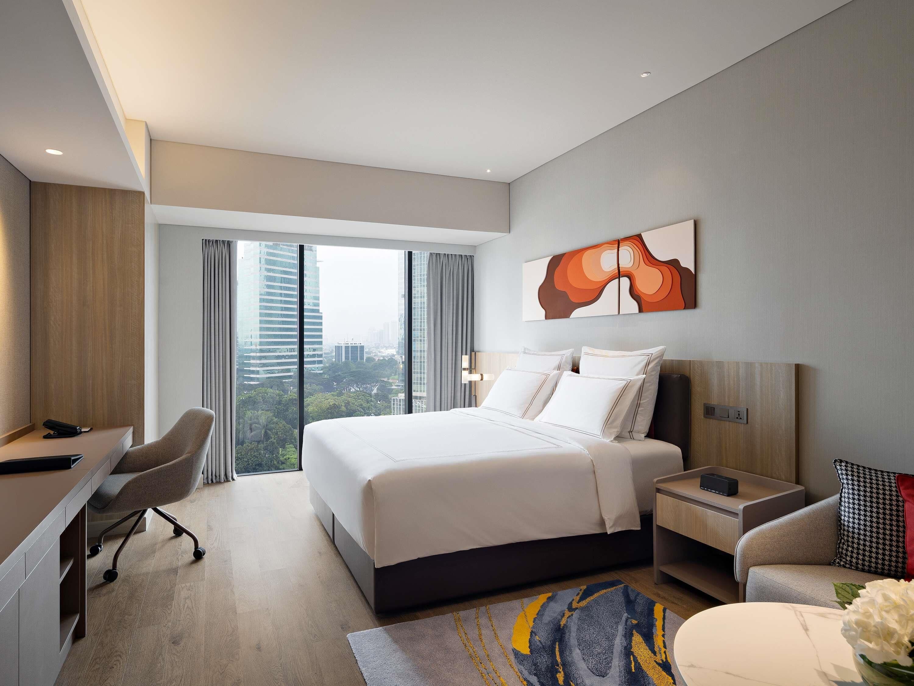 A modern hotel room with a large bed, a desk, and a chair. There is a view of the city through large windows.
