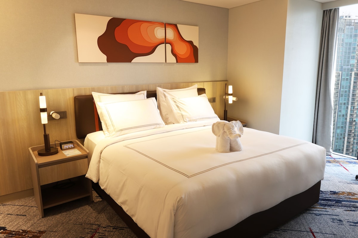 Habitación de hotel con una cama grande, una mesita de noche con un reloj, una obra de arte en la pared y una ventana con vistas a la ciudad.