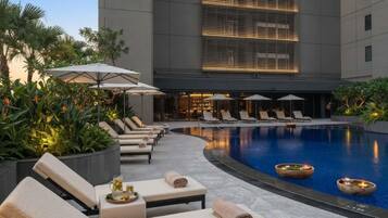 Zona de piscina con sillones, sombrillas y un edificio moderno al fondo.