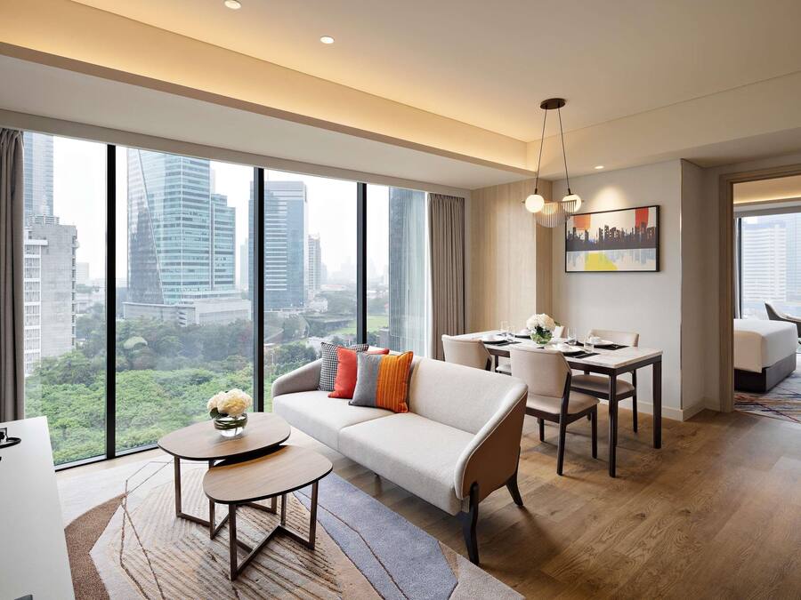 A modern hotel room with a sofa, dining table, and chairs, large windows with a city view, and a painting on the wall.