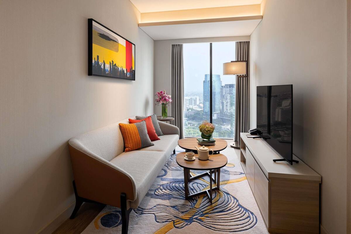 A modern hotel room with a sofa, a small round table with a vase, a television, and a large window with a city view.