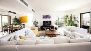 A modern living room with a white sofa, a coffee table, and a TV. There are plants, a dining area, and large windows. - Oceanview Penthouse with Rooftop Plunge Pool & BBQ (Cancún)