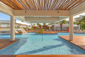 Ein Poolbereich mit klarem Wasser, Holzterrasse, Liegestühlen, Sonnenschirmen und Palmen.