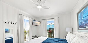 Habitación de hotel con cama, ventilador de techo, televisión, ventana con cortinas y vista a un edificio.