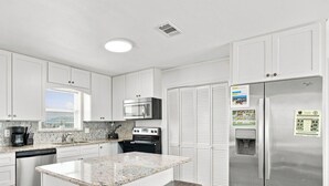Una cocina moderna con armarios blancos, encimera de granito, microondas integrado, refrigerador de acero inoxidable y una ventana con persianas.