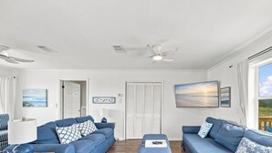 Sala con dos sofás, ventilador de techo, televisor y ventana con vista a una casa.