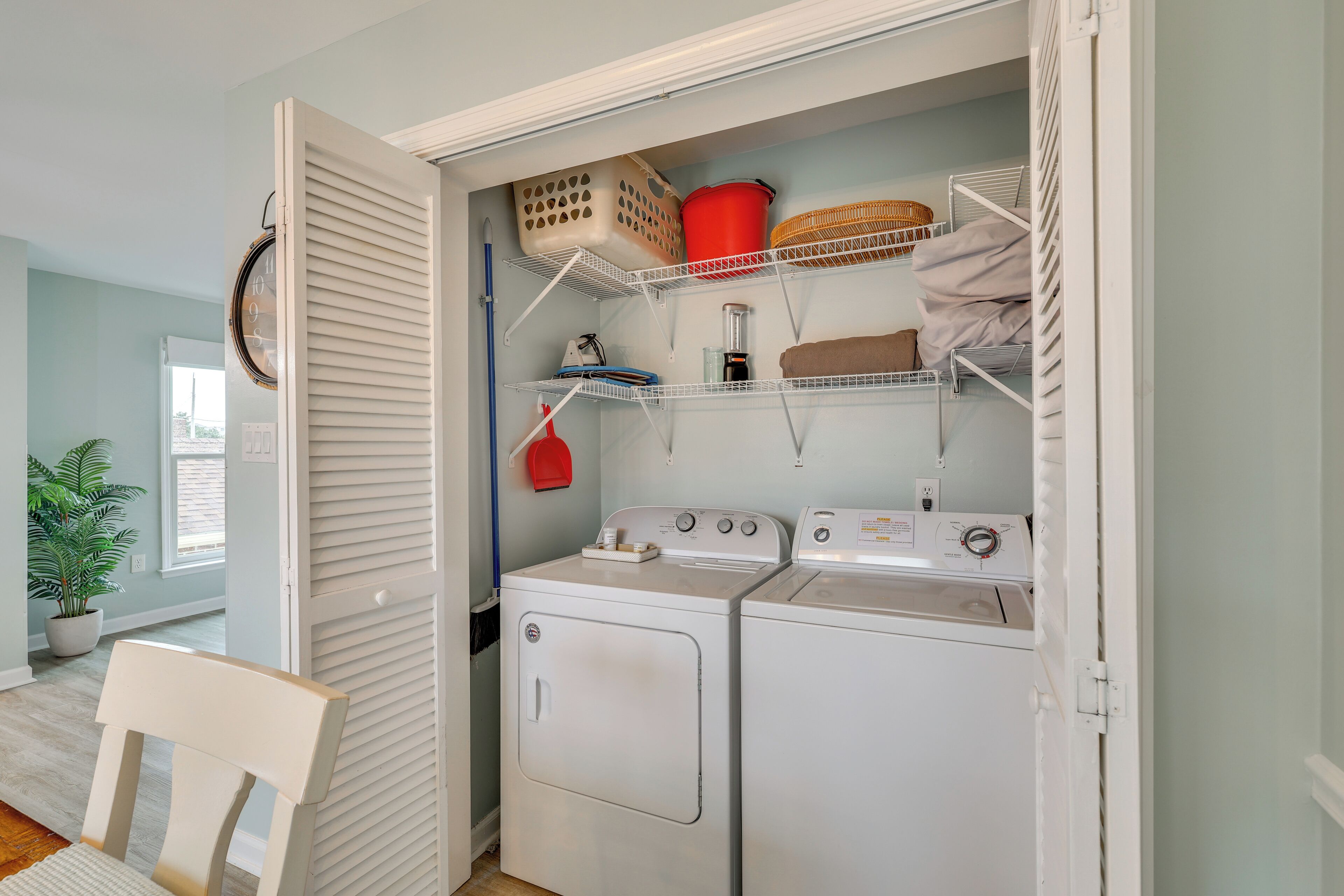 Une buanderie équipée de deux machines à laver et deux sèche-linge blancs, d’étagères avec des paniers et du matériel de lessive, et d’une chaise.