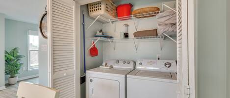 Une buanderie équipée de deux machines à laver et deux sèche-linge blancs, d’étagères avec des paniers et du matériel de lessive, et d’une chaise.