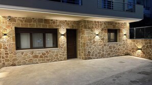 A two-story house with stone exterior walls, a front door, and two windows with exterior lighting. - Casa Nicholas (Malia)