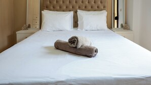 A neatly made bed with white linens and a rolled towel, a tufted headboard, and a nightstand with a mirror and decorative items. - Casa Nicholas (Malia)