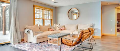 A living room with a beige sofa, a brown leather chair, a patterned rug, and a large window with curtains.