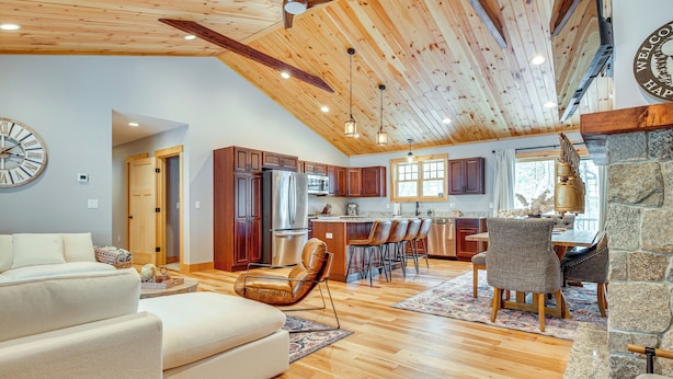 A spacious living room with a large sofa, a dining area with chairs, a kitchen with wooden cabinets, and a stone fireplace. - 6 Mi to Lake Winnipesaukee: New Hampshire Home (Gilford)