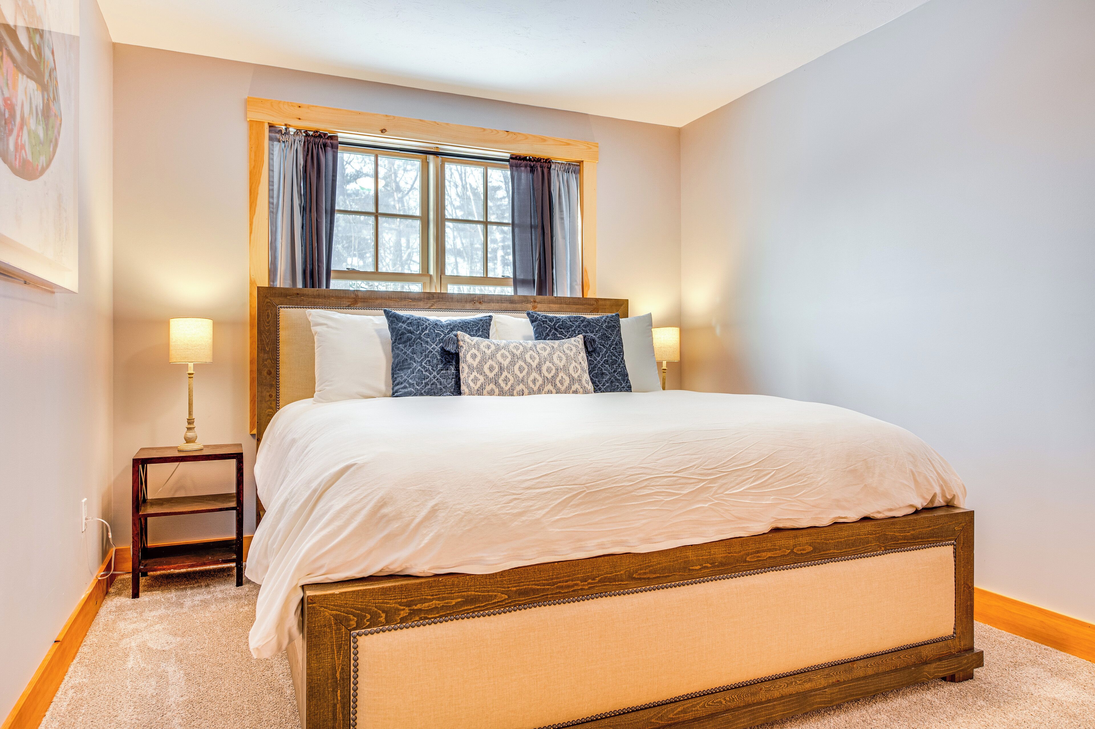 A bedroom with a wooden bed frame, white bedding, a nightstand with a lamp, and a window with curtains.