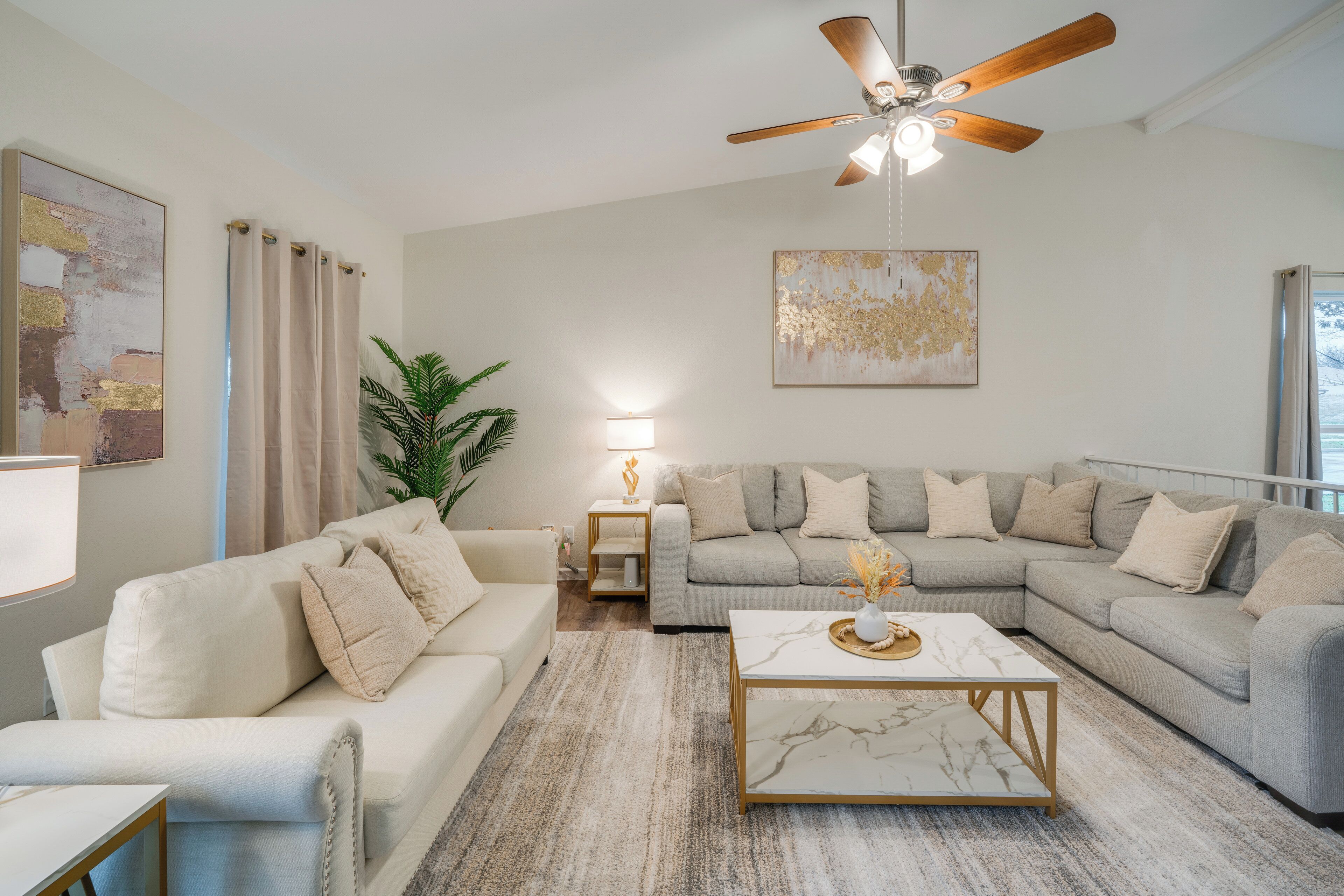 A modern living room with a beige sofa, a white sofa, a marble coffee table, a ceiling fan, and a large abstract painting on the wall.