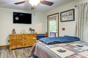 Une chambre à coucher comprenant un lit, une commode en bois, un téléviseur, un ventilateur de plafond et une fenêtre avec des rideaux.
