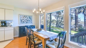 Un coin repas avec une table et des chaises en bois, un lustre, une vue sur les arbres à travers de grandes fenêtres, et une cuisine équipée de placards et d’un radiateur.