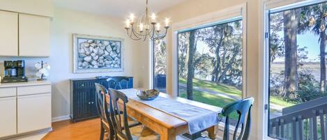 A dining area with a wooden table and chairs, a chandelier, a view of trees through large windows, and a kitchen with cabinets and a radiator.