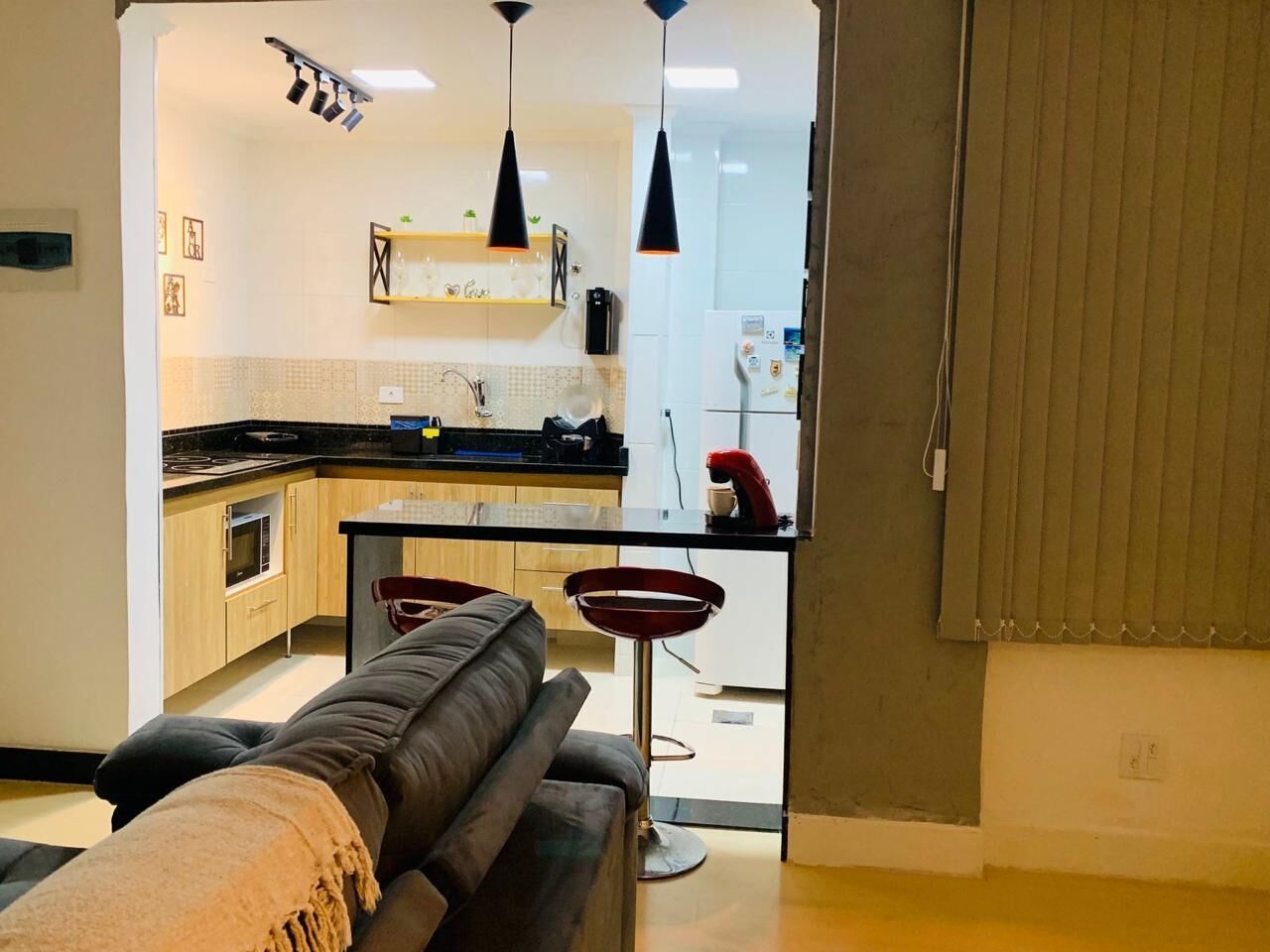 A modern kitchen with black countertops, a built-in oven, and a refrigerator. There is a dining area with a black sofa and two red bar stools.