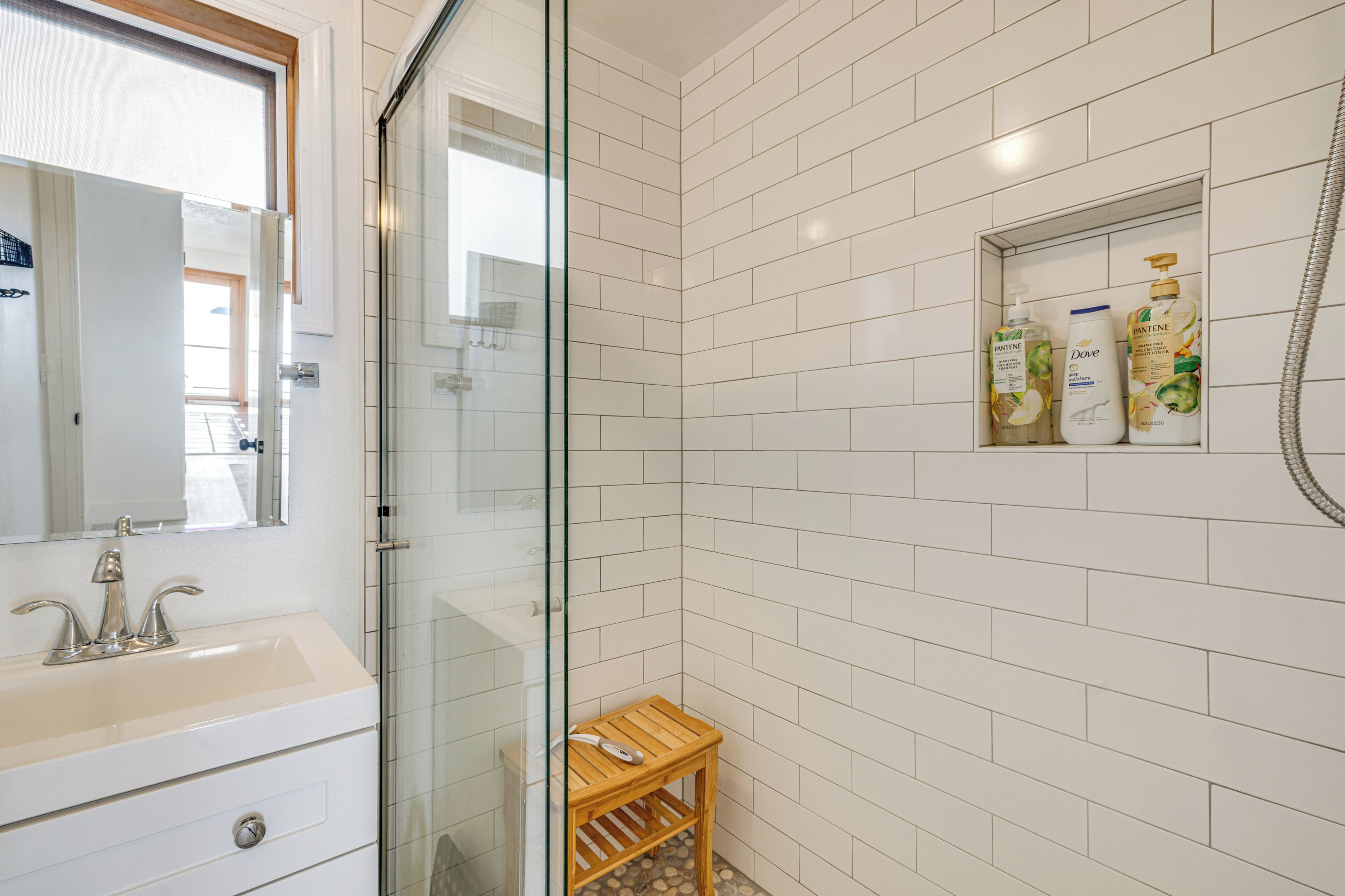 Un baño con paredes de azulejos blancos, cabina de ducha de vidrio, un taburete de madera y un estante con artículos de aseo.