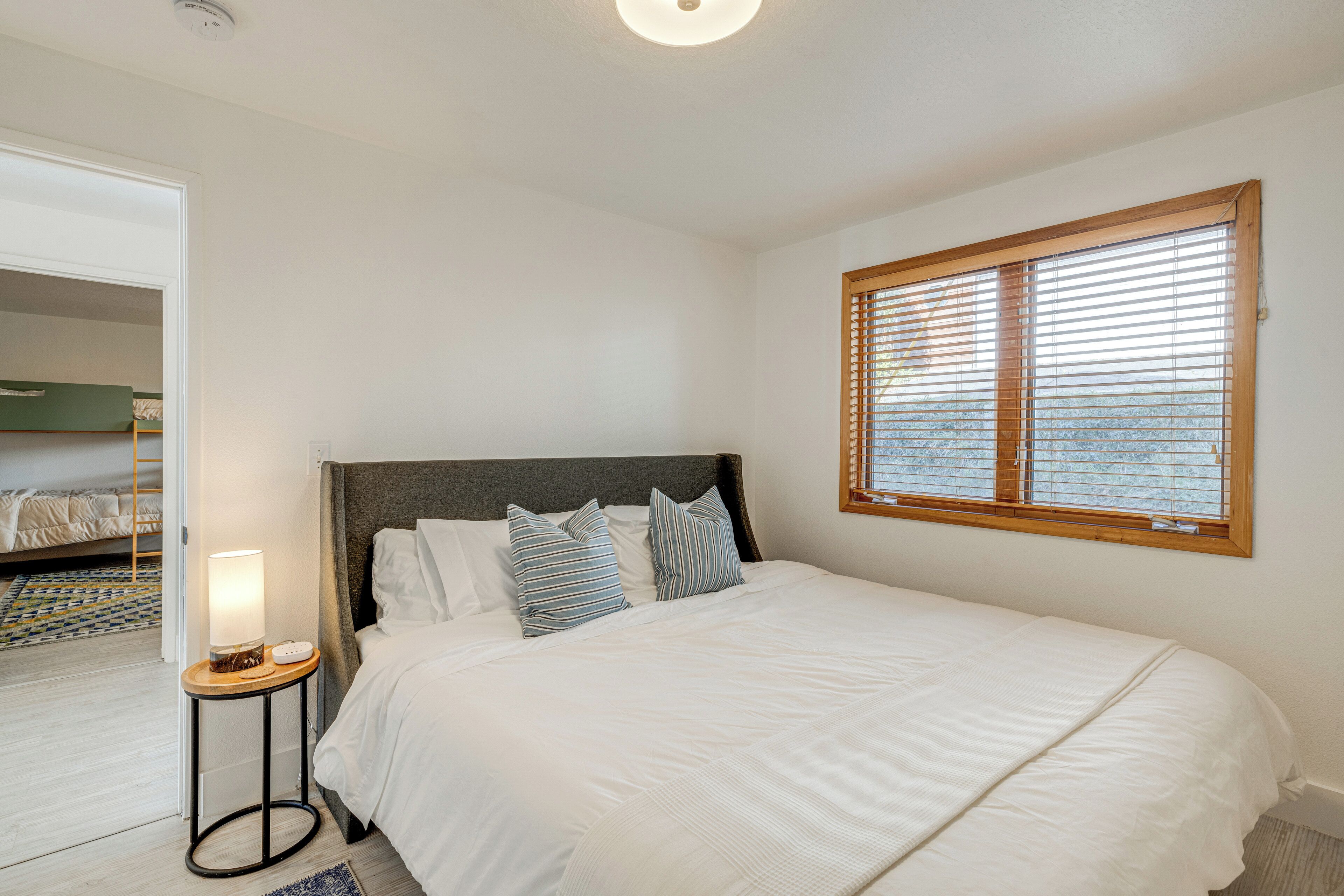A neatly made bed with white bedding and striped pillows, a bedside table with a lamp, and a window with blinds.